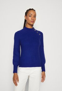Knitted blue sweater with a high neck, ribbed texture, and gold buttons on one shoulder. Fitted design, long sleeves, and a hem that sits at the hips.