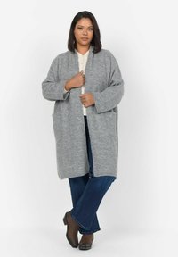 Gilet gris clair en material doux, avec un devant ouvert, une coupe décontractée, deux poches frontales et des manches mi-longues. Associé à un jean évasé.