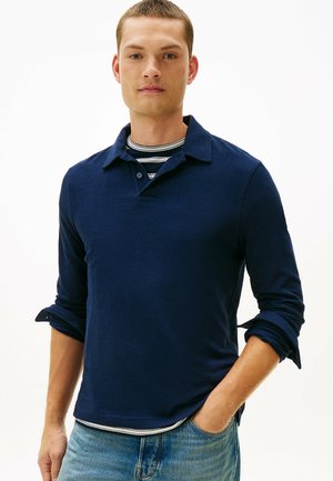 Mand iført en navyblå langærmet polo med op-rullede ærmer over en sort-hvidstribet skjorte, stående med den ene hånd i lommen på lyseblå jeans.