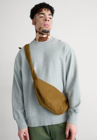 Bolso bandolera marrón hecho de tela suave, con una forma triangular única y una sola correa para el hombro, llevado con un sudadera gris.