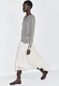 Szary top z długimi rękawami, biała zwiewna spódnica, brązowe botki w stylu slouchy i srebrna bransoleta noszona na nadgarstku. Gładkie tekstury i minimalistyczny design.