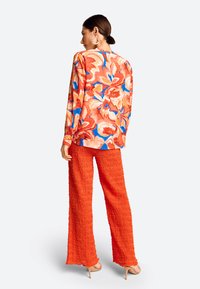 Mehrfarbige Blumenbluse, orangefarbene, strukturierte weite Hosen und beigefarbene High Heels. Die Bluse hat ein lebendiges Muster mit blauen und pinken Akzenten.