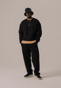 Schwarzer Fleece-Hoodie mit einem beigen gerippten Saum, kombiniert mit passenden Hosen und einem schwarzen Bucket Hat. Goldenes Logo-Detail auf der Brust.