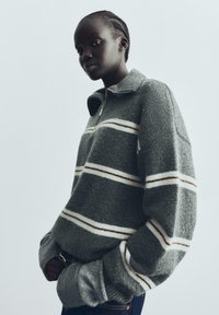 Sivi flis pulover s bijelim i smeškim horizontalnim prugama, kravatom na pola zatvarača i oversized rukavima. Mekana tekstura i opušten kroj.