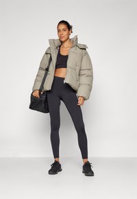 Napuhao svijetlosivi kaput s visokim ovratnikom, crni crop top, crne tajice i crne tenisice; drži crnu torbu s trakama.