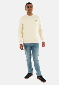 Pull crema con polsini a coste, abbinato a jeans azzurri chiari e sneakers nere. Il pullover presenta un piccolo logo nero sul petto.