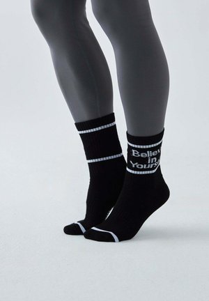 Osoba nosząca czarne skarpetki z białymi paskami i napisem „Uwierz w siebie” na jednej skarpecie, stojąca na palcach na jednolitym tle.