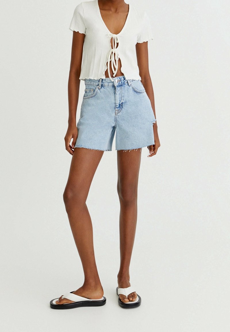 Shorts en denim bleu clair avec bords effilochés, associés à un crop top côtelé blanc avec un décolleté en V et un nœud à l'avant. Chaussures : tongs blanches.