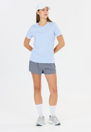 Femme portant un t-shirt bleu clair, un short gris, une casquette blanche, des chaussettes blanches et des chaussures de course grises, debout les mains derrière le dos, regardant vers la droite.
