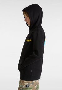 Felpa nera con cappuccio e tasca frontale, decorata con un grande logo giallo "VANS". Presenta un cappuccio regolabile e viene indossata sopra pantaloni mimetici.