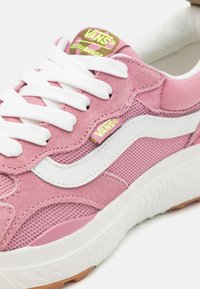 A rózsaszín Vans sportcipők velúr és hálós anyagból készültek, fehér bőr részletekkel, vastag fehér fűzőkkel és texturált gumi talppal rendelkeznek.