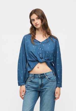 Młoda kobieta w niebieskiej, długiej, zapinanej na guziki koszuli skręconej w talii i niebieskich dżinsach, stojąca na jednolitym tle.