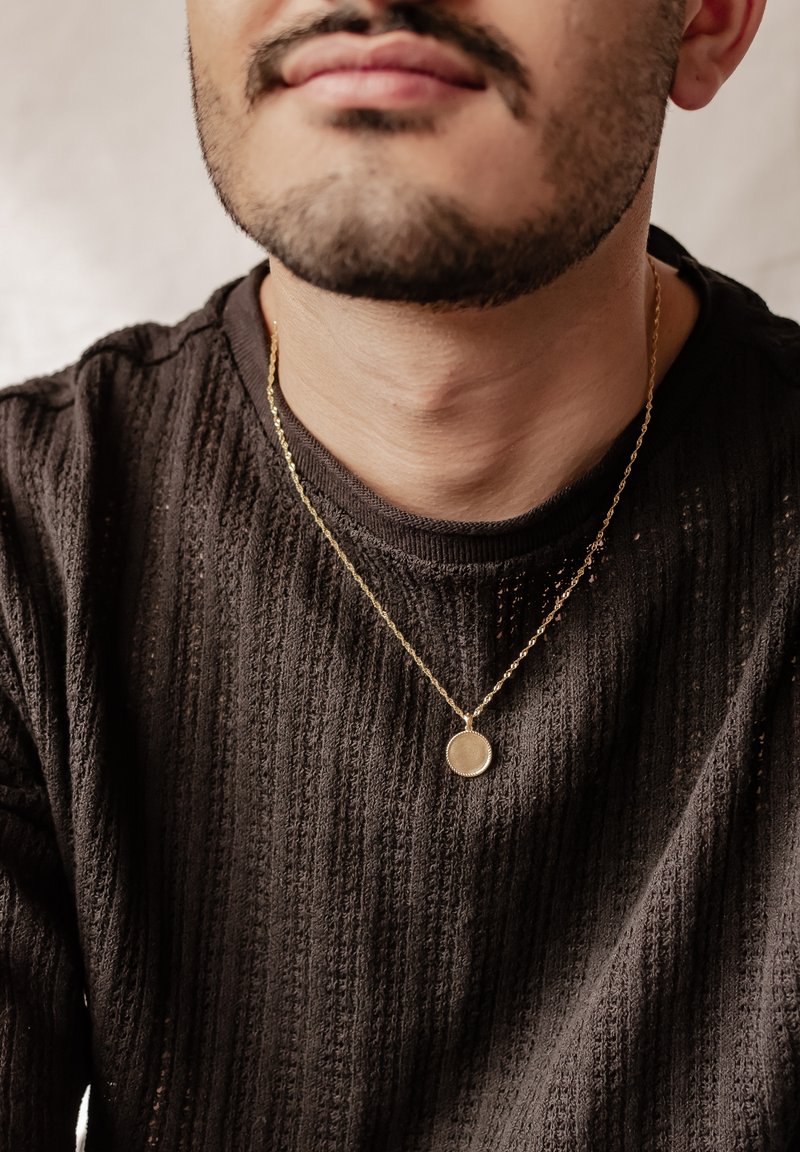 Collana dorata con pendente circolare, indossata sopra un maglione scuro con texture e un motivo a coste, dal collo rotondo.