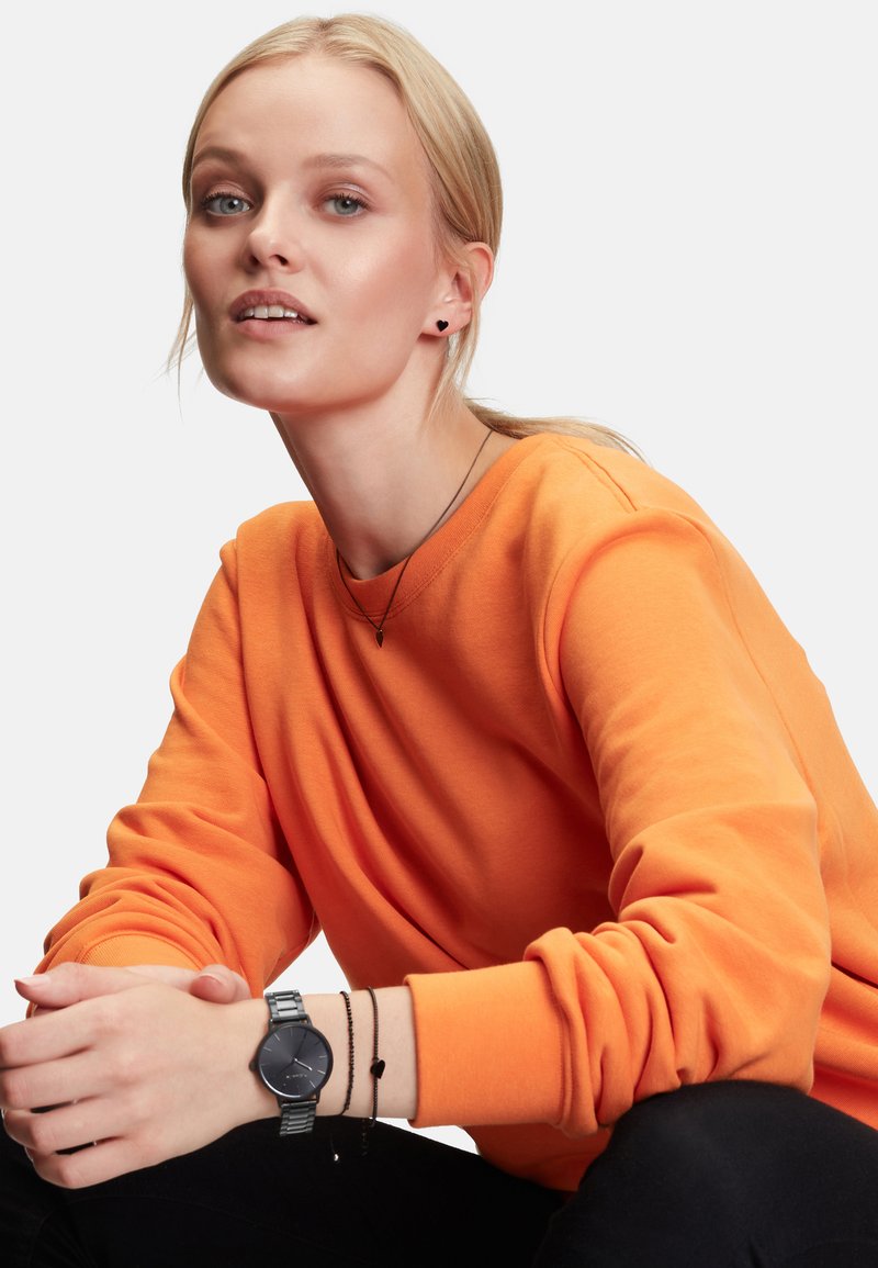 Zwarte horloge met een ronde wijzerplaat en metalen band. Model draagt een oranje sweater, gelaagd met een dunne zwarte ketting en armband.