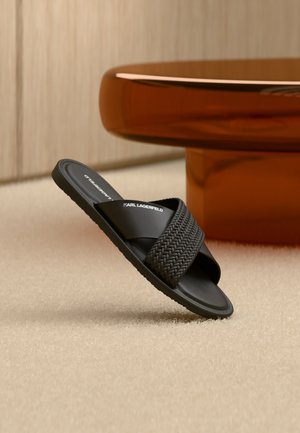 Zwarte slidesandaal met gevlochten band en "KARL LAGERFELD" logo, geplaatst op een beige tapijt naast een ronde amberkleurige tafel.