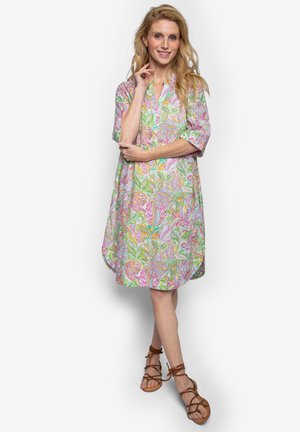 Kobieta o długich blond włosach, ubrana w kolorową, kwiecistą sukienkę do kolan i brązowe sandały na sznurowadła, stoi ze skrzyżowanymi ramionami i uśmiecha się.