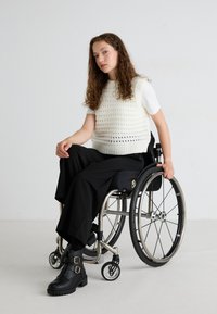 Gilet bianco a maglia sopra una maglietta bianca aderente, abbinato a pantaloni neri a gamba larga e stivali neri chunky alla caviglia. Utente in sedia a rotelle.