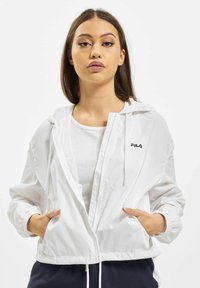 Valkoinen kevyt vetoketjullinen takki hupulla, joustavilla hihansuilla ja kahdella sivutaskulla. Rinnassa on Fila-logo. Tyylitelty valkoisen paidan päällä.