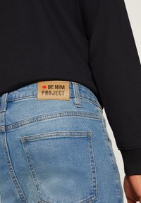 Világos mosású farmernadrág barna bőrcímkével, minimalista dizájnnal és jellegzetes varrással, oldalról, fekete felső ellen.