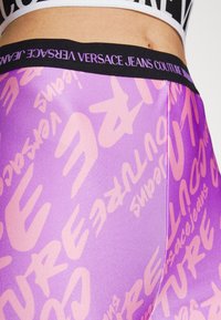 Lila leggings med ett upprepat rosa logotryck och ett svart elastiskt midjeband med "Versace Jeans Couture" präglat i vit text.