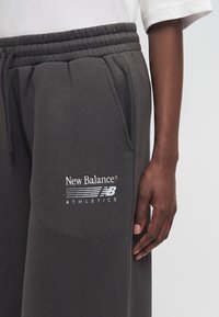 Άτομο φορεί σκούρο γκρι φόρμα New Balance με ελαστική μέση και πλαϊνή τσέπη, καθώς και ένα λευκό κοντομάνικο πουκάμισο.