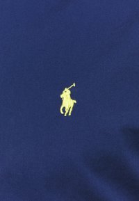 Tmavě modrá látka s hladkým povrchem, na levé straně s malým žlutým vyšívaným logem koně a jezdce v postoji pro pólo.