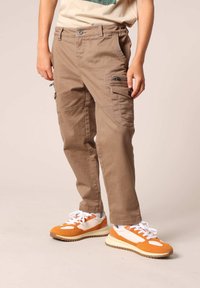 Khaki cargobroeken met meerdere zakken, gestructureerde stof en een comfortabele pasvorm, gecombineerd met oranje en witte sneakers, maken de look compleet.