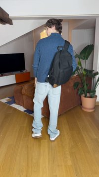Black backpack, knitted blue sweater, light blue jeans. Indoor setting with a brown couch, TV, plants, and wooden flooring.