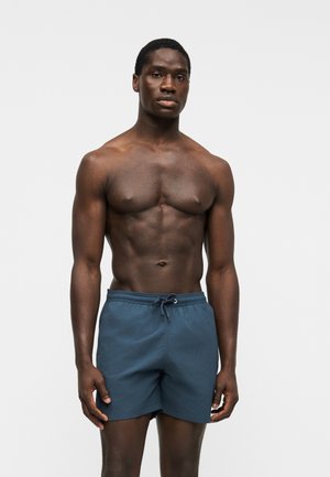Homme musclé à la peau foncée, debout face à l'objectif, portant un short de bain bleu avec cordon, sur un fond blanc uni.