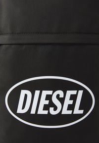Juodas maišelis pagamintas iš medžiagos, su išskirtiniu baltu "DIESEL" logotipu ovalo formos dizainu, su lygia tekstūra ir matomomis siūlų akcentais.