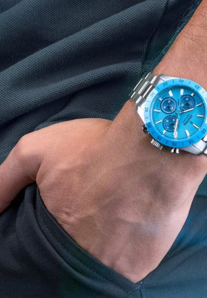 Polso con orologio in acciaio inossidabile con quadrante e lunetta di un brillante blu, tre quadranti secondari e finestra della data, appoggiato su tessuto scuro e strutturato.