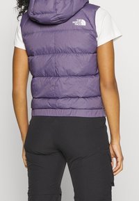 Fioletowa puchowa kamizelka z kapturem i białym logo, z pikowanymi panelami i dopasowaną talią, w połączeniu z czarnymi spodniami cargo.