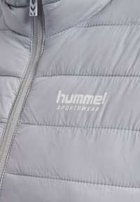 Lys grå pufferjakke med vandret syninger, lynlås foran og et hvidt "hummel SPORTSWEAR" logo iøjnefaldende placeret.