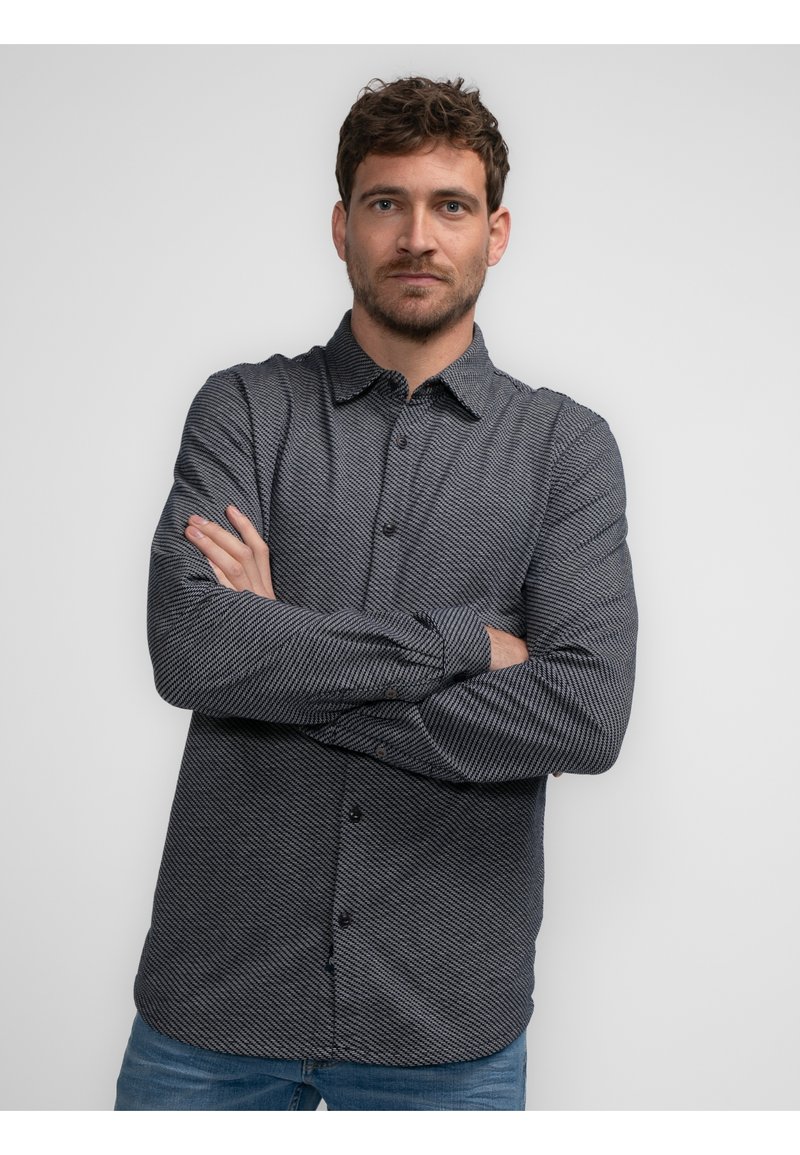 Stribet sort-hvid skjorte med knaplukning og krave, lange ærmer og lige kant. Model iført tætsiddende denim jeans med armene korslagte.