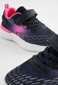 Baskets avec un dessus en mesh à motifs bleu foncé et violet, accents noirs, doublure intérieure rose, et une sangle Velcro sur des lacets traditionnels. Semelle blanche.