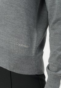 Szürke kötött pulóver bordás mandzsettával és aljjal. Diszkrét "Calvin Klein" logó hímzett a bal alsó oldalán, puha tapintású és testesített fazonú.