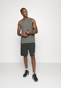 Grå, ermeløs treningstopp, svarte shorts med logo og svarte joggesko. Modellen holder en vannflaske og står mot en ensfarget bakgrunn.