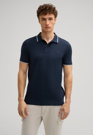Marineblaues Polohemd mit klassischem Kragen, weißen Akzentstreifen am Kragen und den Ärmeln sowie einem dezenten Logo auf der Brust. Glatte Stofftextur.