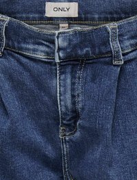 Jeans de denim en azul medio con una textura suave. Cuentan con un cierre de cremallera, pasadores para el cinturón y una pequeña etiqueta de marca en la cinturilla.