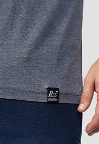 Šedé tričko s jemnou texturou, na spodní části s černou etikou označenou "R:C RE-COVERED". Ruka odpočívající blízko švu.