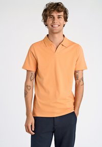 Polo arancione con un collo corto e maniche corte, realizzato in un tessuto liscio. Indossato con pantaloni blu navy, che mettono in mostra tatuaggi visibili sulle braccia.