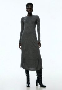 Graues langärmliges Kleid aus einem weichen Stoff, mit einer figurschmeichelnden Silhouette, rundem Ausschnitt und einem seitlichen Schlitz, kombiniert mit schwarzen, kniehohen Stiefeln.