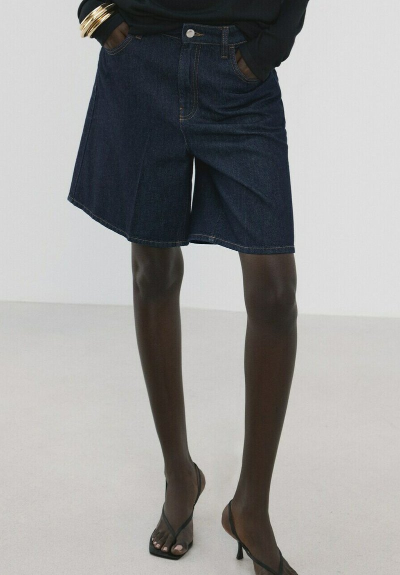Personne portant un short en denim foncé jusqu'aux genoux, des talons noirs à brides, et un haut à manches longues noir avec des bracelets dorés, debout les mains dans les poches.