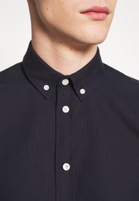 Camisa azul oscura de botonadura clásica, con un cuello clásico, presentada con botones blancos y una tela suave y texturada. Vista en primer plano del escote.