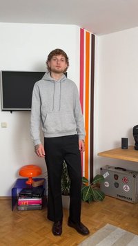 Sweatshirt cinza com capuz e bolso frontal, combinado com calças pretas. O cenário inclui listras coloridas na parede e elementos decorativos.