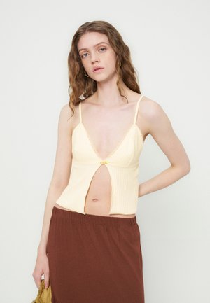 Jeune femme aux cheveux ondulés porte un haut sans manches jaune pâle avec une fente à l'avant et une jupe marron, debout devant un fond uni.