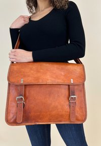 Bruine leren boodschappentas met een klep, voorzien van zilverkleurige metalen accenten en twee verstelbare riemen. Gladde textuur en een licht verweerde afwerking.