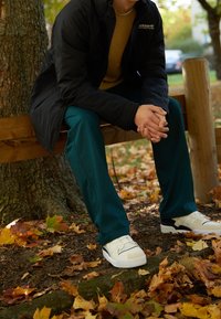 Gröna lätta byxor, svart jacka och beige sweatshirt i kombination med vita sneakers. Omgiven av höstlöv och sittande på en bänk av trä.