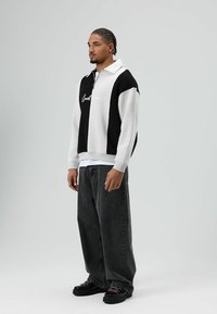 Zwart-witte pullover met ritskraag, voorzien van een brede streep en geborduurde tekst. Gecombineerd met losse donkergroene spijkerbroek en zwarte schoenen.