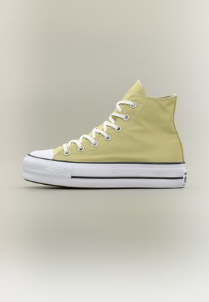 Zapatilla de lona alta en amarillo pálido con suela y puntera de goma blancas, cordones blancos y detalle de raya negra a lo largo de la suela.